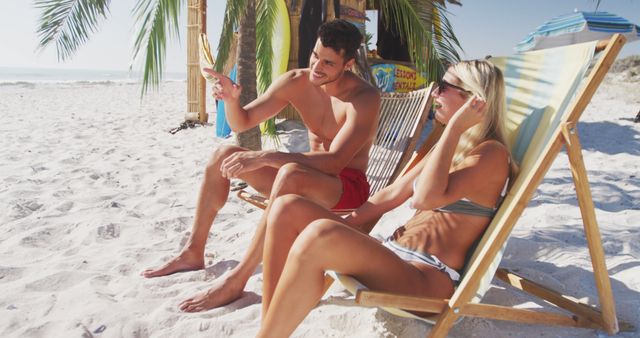 Couple is relaxing on a sandy beach under palm trees. They are sunbathing in swimwear, with the ocean visible in the background. Perfect for websites or advertisements related to summer vacations, tropical getaways, travel destinations, and relaxation.