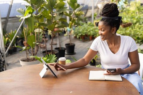 African american woman working on tablet and phone in greenhouse nursery with notebook