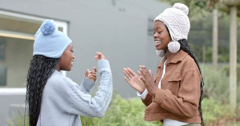 Two african american girls playing clap game outdoors wearing pom-pom hats and sweaters