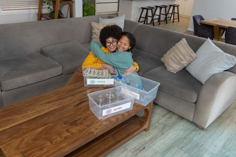 African american mother hugging child on sofa while sorting paper and glass recycling bins