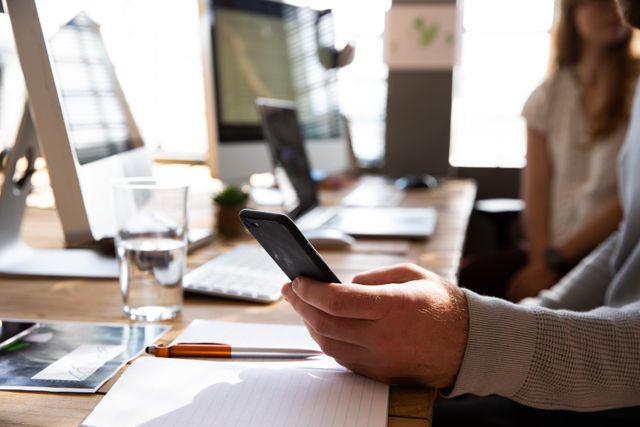 Side view mid section of Caucasian creative professional man in a modern office, holding in hand a smartphone, sitting at the desk, in the background his colleague working at her desk, out of focus.