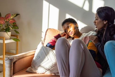 Black woman looking pensive while south asian friend comforting on sunlit cozy sofa