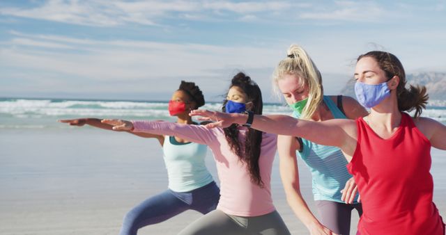Group of diverse female friends wearing face masks practicing yoga at the beach. healthy active lifestyle, outdoor fitness and wellbeing.