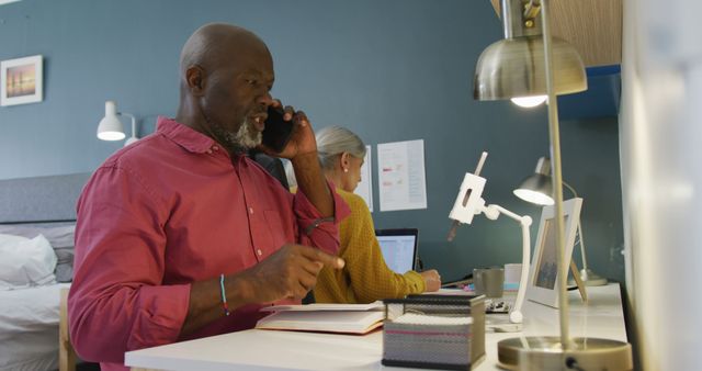 Elderly couple working from home office, engaged in various tasks. Man having a phone conversation while woman using laptop in background. Perfect for content on remote work, productivity, elderly lifestyle, home workspace setup, and work-life balance.