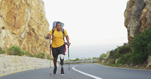 Determined biracial man with prosthetic leg trekking with backpack and walking poles on coastal road. Long distance walking, fitness, challenge, disability and healthy outdoor lifestyle.