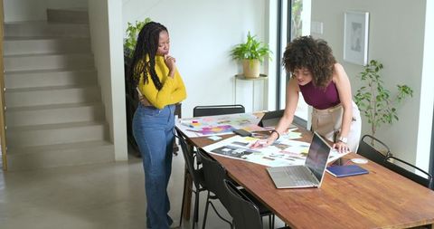 Diverse female colleagues collaborating at modern table with laptop and design moodboards