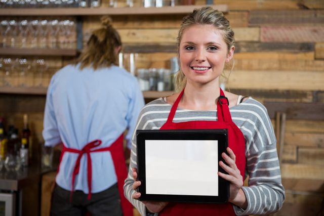 Portrait of smiling young woman showing digital tablet with male colleague in background at coffee shop