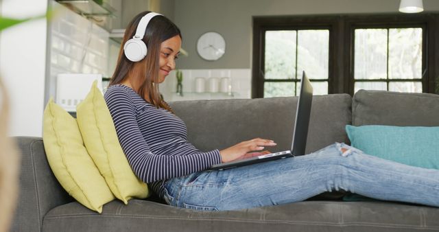 Biracial teenager girl sitting on sofa, using laptop and headphones. Spending quality time, lifestyle and adolescence concept.