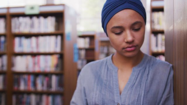 Young woman wearing blue hijab is reading peacefully in a library environment. Elevated focus on studying and education. Ideal for illustrating concepts related to learning, cultural inclusion, or academic articles. Suitable for talk about library resources or highlighting educational diversity.