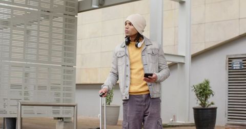 Urban traveler standing with suitcase and smartphone wearing beanie and headphones