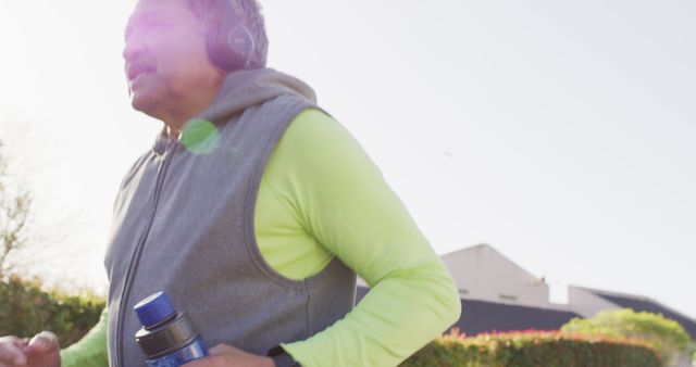 Elderly man taking care of his health by jogging outdoors on a sunny day. Wearing a gray vest and neon green long sleeve, he enjoys his run with headphones and carries a water bottle. Ideal for use in articles about senior fitness, healthcare, active lifestyles, and wellbeing.