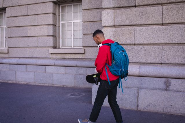 Young man with a backpack and skateboard walking in an urban area. Ideal for use in travel blogs, lifestyle magazines, or advertisements promoting city exploration, youth culture, and outdoor activities.