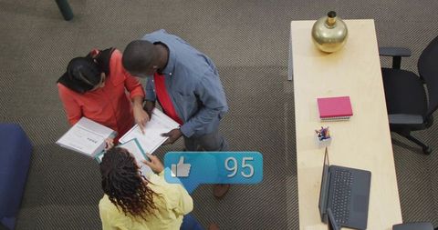 Top-down office team collaborating over documents and laptop with social likes overlay