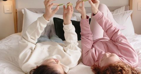 Friends relaxing on bed in cozy loungewear holding green fruit slices and smiling
