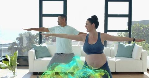 Pregnant woman practicing prenatal yoga with partner, balancing and smiling in sunlit living room