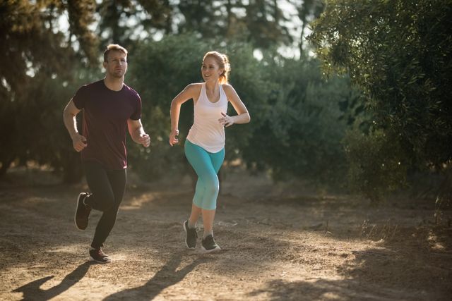 Young couple running together outdoors on a sunny day, surrounded by nature and trees. Ideal for promoting fitness, healthy lifestyle, outdoor activities, and exercise routines. Suitable for use in advertisements, fitness blogs, health and wellness websites, and social media campaigns.