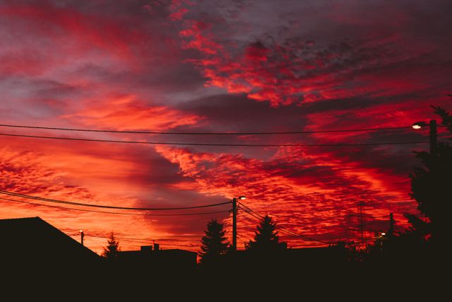 Capturing a breathtaking red sunset over an urban landscape, this scene shows a silhouette of power lines, houses, and trees against a strikingly vibrant sky filled with dramatic clouds. Perfect for use in cityscape themes, nature photography collections, travel blog headers, background elements for presentations, or artistic projects focused on evening and twilight settings.