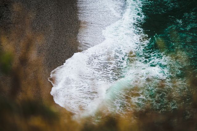 A close-up aerial view of ocean waves gently washing over a pebble beach. This image is perfect for coastal themes, travel promotions, and marine-related content. The contrasting textures of the pebbles and waves give a peaceful yet dynamic feel, making it suitable for backgrounds, posters, or website banners focused on nature and the environment.