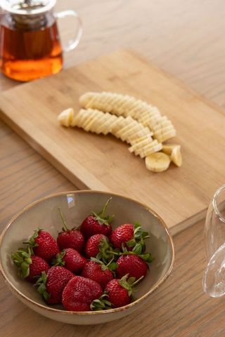 Rustic ceramic bowl holding strawberries on wooden table with sliced banana and glass teapot