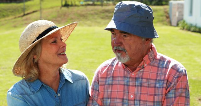 A Caucasian senior couple enjoys a sunny day outdoors, wearing casual hats and smiling at each other, with copy space. Their warm interaction suggests a moment of leisure and companionship in a serene environment.