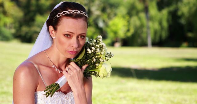 Bride in wedding dress holding bouquet of flowers alone in an outdoor garden. She looks sad and contemplative. Ideal for use in articles about bridal emotions, wedding photography, and relationship struggles. Suitable for wedding services marketing, mental health resources, and dramatic narratives.