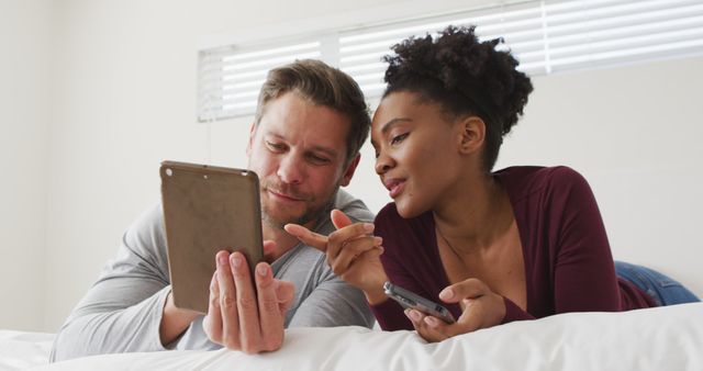 Couple is lying on bed while enjoying their electronic devices. Man is holding a tablet and woman is holding a smartphone, both smiling and interacting with each other. Perfect for depicting a technology-friendly lifestyle, intimate moments shared in a home setting, or marketing for tech products and home relaxation.