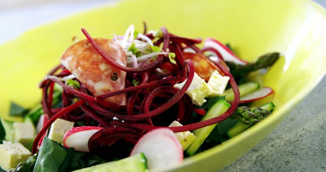 This vibrant salad features an array of fresh vegetables like spiral beets, radishes, asparagus, and cucumbers, topped with cubes of feta cheese and microgreens in a yellow bowl. Ideal for promoting healthy food choices, restaurant menus, food blogs, and culinary magazines showcasing nutritious meal options.