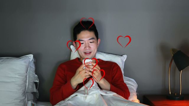 Asian man is lying in bed, smiling while using his smartphone. His face is slightly illuminated by the screen's glow, while multiple red heart icons are animated around him, suggesting love or positive emotions. It is well-suited for use in topics related to online dating, social media interaction, or expressing feelings through technology. This video could also be used in blogs about digital love, late-night texting, or connecting through technology.