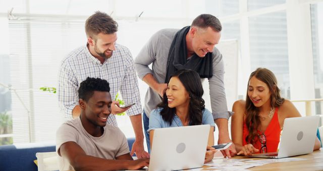 Diverse group of professionals in modern office collaborate on project, using laptops and documents. Team members from different backgrounds interact positively, discussing ideas and strategies. Useful for corporate content, teamwork themes, diversity in workplace promotions, collaborative project visuals.