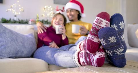 Cozy couple relaxing on sofa in festive knit socks and santa hat holding warm mugs