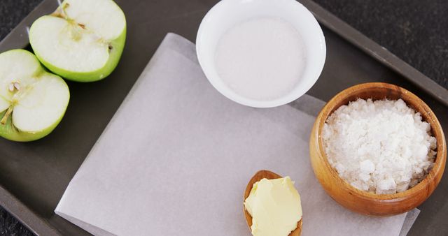 Fresh ingredients on a baking tray: green apple halved, butter, flour, and sugar in individual bowls. Ideal for food blogs, recipe websites, cooking guides, and baking tutorials.