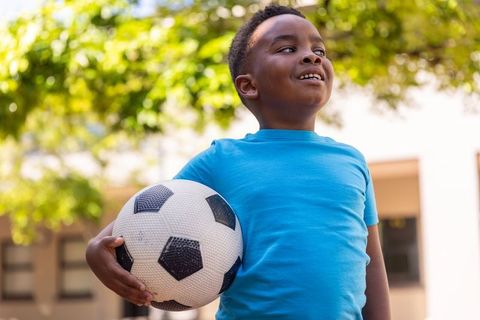 Confident african american boy holding soccer ball and smiling in sunny schoolyard
