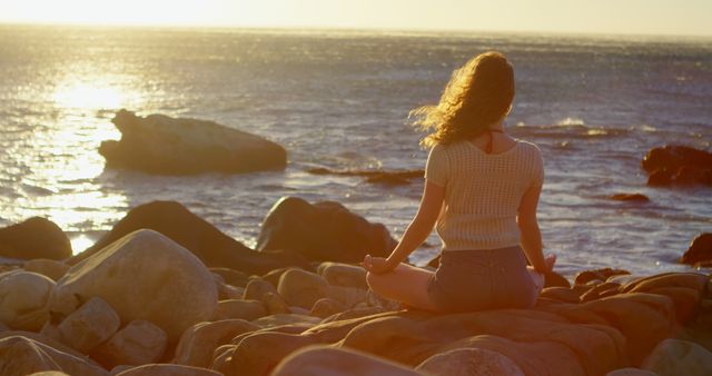 Young Caucasian woman meditates on a rocky beach at sunset. She finds tranquility in the serene outdoor setting as waves gently crash nearby.