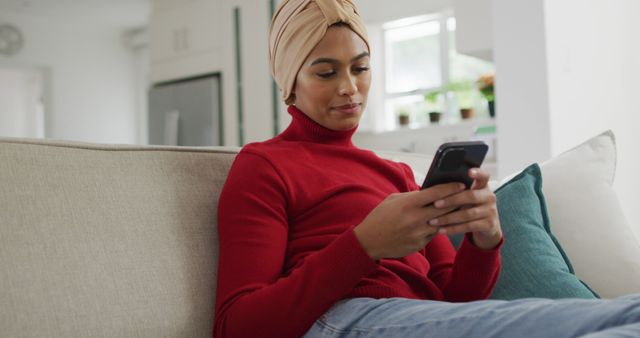Image of happy biracial woman in hijab sitting on sofa and using smartphone. Lifestyle, spending free time at home with technology concept.