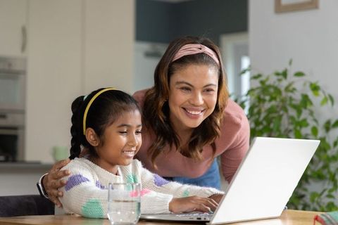 Asian mother guiding daughter on laptop at kitchen table, smiling and learning together
