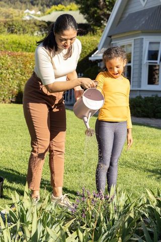 Multiracial mother and daughter watering front-yard flowerbed with pink watering can