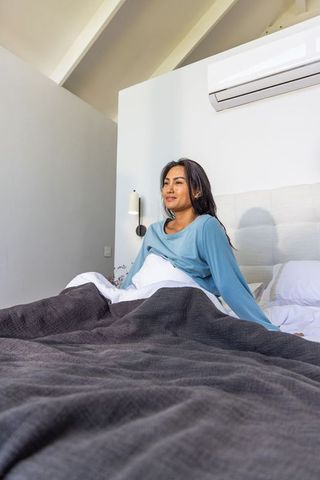 Asian woman sitting up in bright modern bedroom wearing light blue loungewear enjoying calm morning