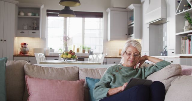 Elderly woman sitting comfortably on a sofa in a home setting, reading or browsing on a digital tablet. Modern kitchen visible in background. Perfect for promoting digital literacy among seniors, elder lifestyle content, home comfort products or smart home tech.