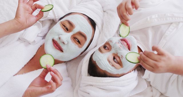 Mother and daughter are lounging in bathrobes with facial masks and holding cucumber slices over their eyes. This image depicts a peaceful and bonding moment. Ideal for use in promotional materials for spa services, skincare products, family wellness programs, and self-care campaigns.