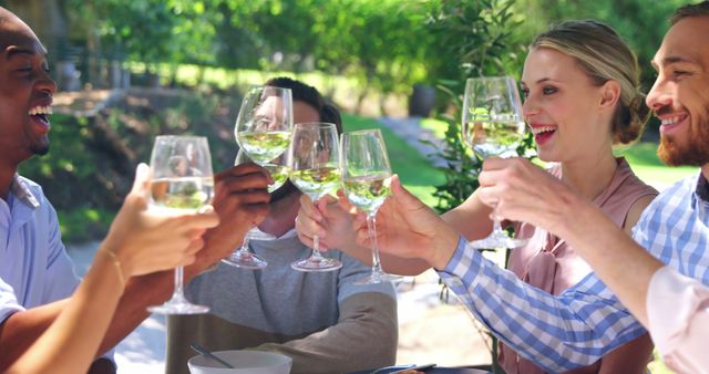 Group of adult friends smiling and toasting with glasses of white wine at an outdoor setting, possibly in a garden or park. The atmosphere appears relaxed, joyful, and celebratory, making it suitable for use in advertisements and articles about social gatherings, outdoor activities, summer events, and friends spending quality time together. Ideal for conveying themes of friendship, celebration, and enjoyment.