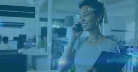 Smiling professional talking on phone while holding clipboard in modern office tech hub