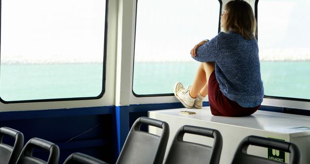 A girl sits alone on a ferry staring out at the sea through the large windows. She appears thoughtful and in contemplation, resting her feet on the bench, suggesting a peaceful, introspective moment. This image can be used to depict themes of travel, solitude, reflection, and peaceful moments. Ideal for articles about travel experiences, personal reflection, or ocean journeys.