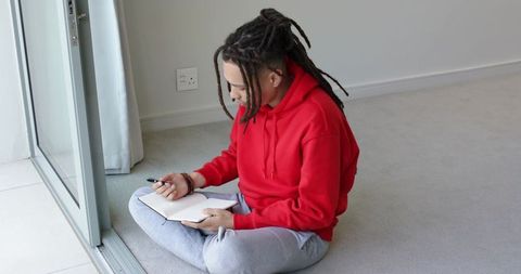 African-american man journaling on floor by sliding glass door in red hoodie, modern home