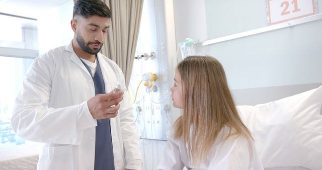 Diverse male doctor showing girl patient in hospital bed how to use asthma inhaler. Consultation, treatment, childhood, illness, medical services, healthcare and hospital, unaltered.