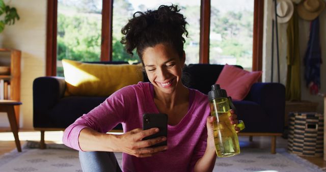 Woman smiling indoors holding smartphone and water bottle, sitting on floor in bright living room. Ideal for content related to lifestyle, relaxation at home, technology use, and healthy hydration.