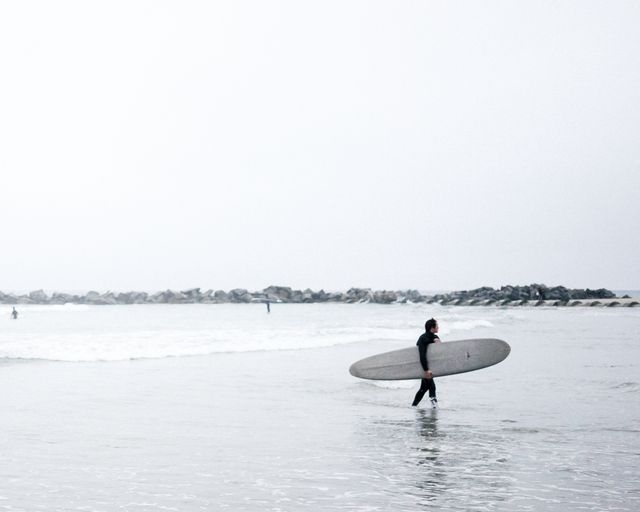 Surfer walks through shallow water carrying surfboard on an overcast day. Rocks and waves seen in the distance. Ideal for beach, surfing, adventure travel, water sports, lifestyle, or summer vacation themes.