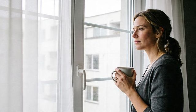 Woman holding mug and gazing out window in apartment, quiet morning urban lifestyle