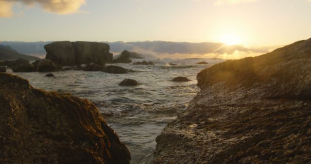Sunset over rocky shore depicted with waves crashing against rocks, creating a serene coastal scene perfect for use in travel promotions, nature-themed projects, and relaxation material presentations.