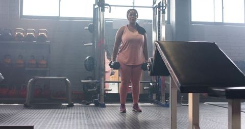 Mid-adult african american woman lifting dumbbells in pink workout set backlit gym strength