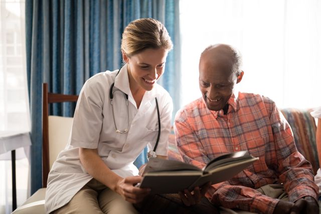 Smiling female doctor reading book with senior man sitting on furniture against window at retirement home